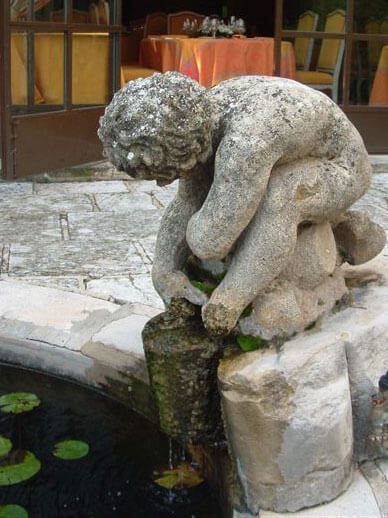 Statue  en pierre d'un personnage accroupi haut-dessus du jet d'eau d'une fontaine.