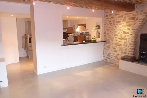 Salon-cuisine, murs en pierre et cheminée sur sol béton ciré clair.
